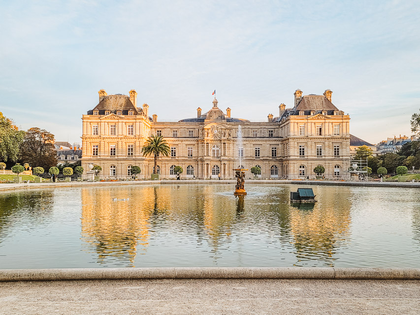 The Palais de Luxembourg, with its off-white walls and black room stands behind the main basin and fountain in the foreground, in the jardin de luxembourg. 