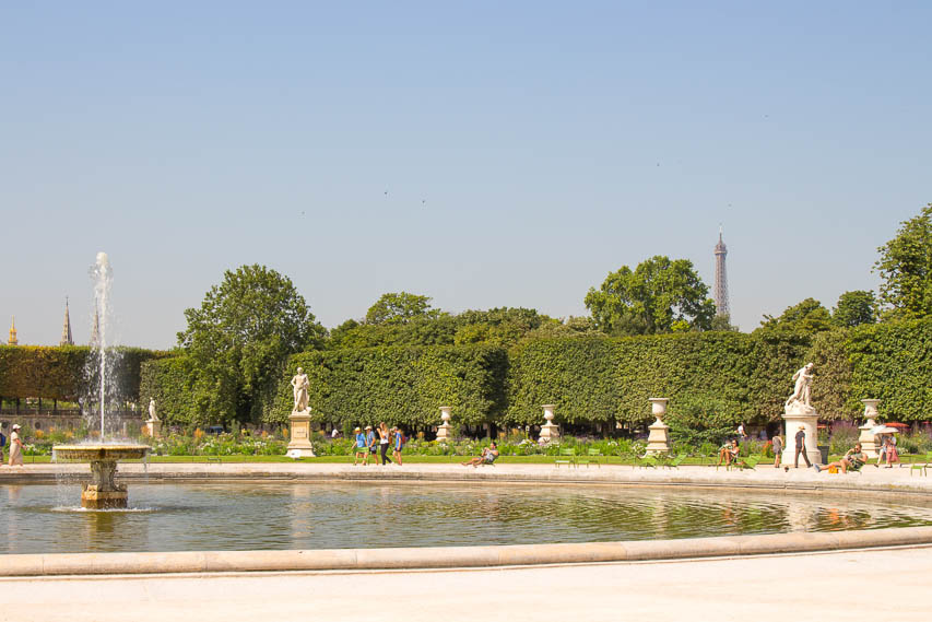 A fountain in a large basin, surrounded by pathways with chairs, sculptures, and green hedges behind it. You can see the top of the Eiffel Tower peaking out above the hedges. 