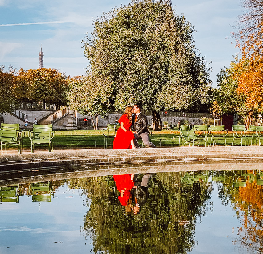 A woman in a red dress and a man kiss while sitting on chairs in front of a basin in the Jardin de Tuileries. The Eiffel Tower is in the background. 