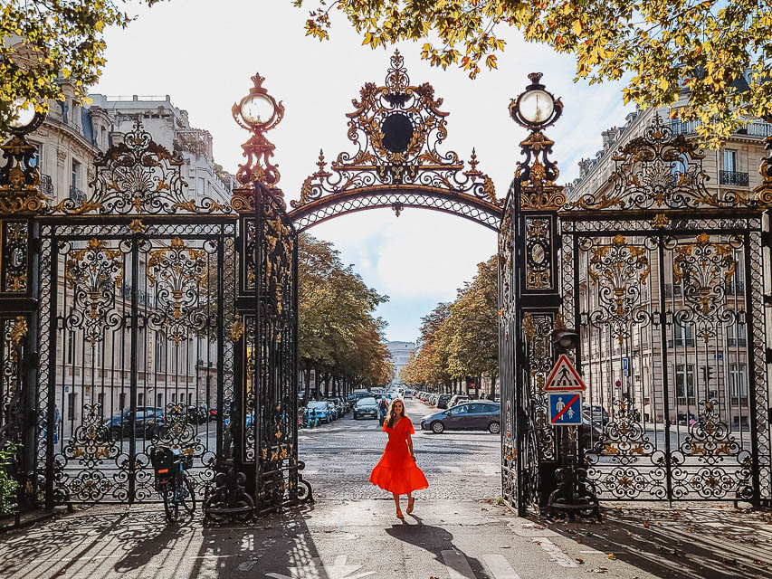 A woman in a red dress stands in the middle of a tall, wrought iron gate with fancy, told-tipped decorations. The streets of Paris and the Arc de Trioimphe are visible behind her. 