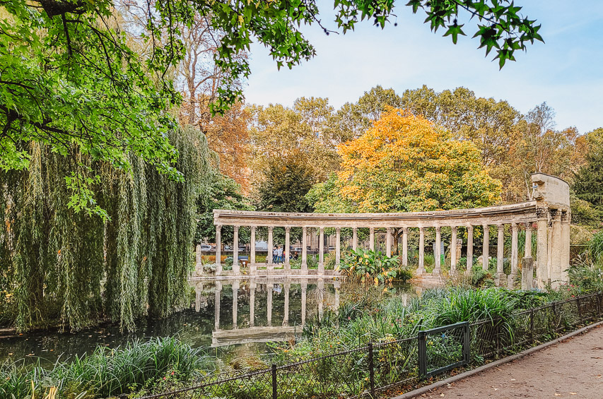 A curved line of columns sits in a pond by a weeping wiloows, with trees in the background. 