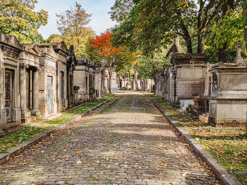 A cobblestoned path goes through a tree-filled cemetery, with old, historic, fairly large above ground tombs line the pathway. 