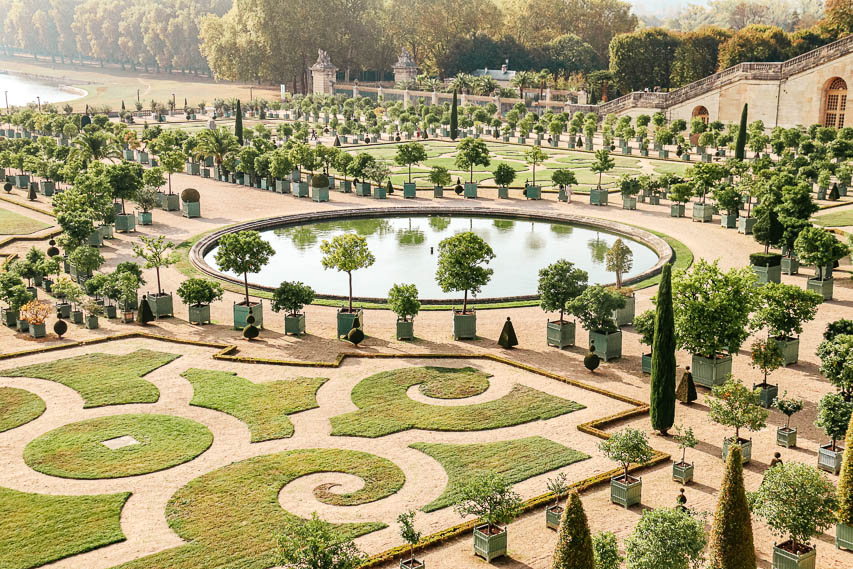 the manicured, heavily landscaped gardens of Versailes, one of the most beautiful gardens in Paris. There's a basin in the middle, curlicue designs in the hedges and grounds, and plants and trees everywhere. 