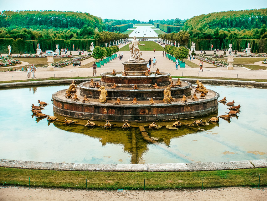 The large fountain in Versailles, with a long, wide pathway behind it, lined with sculputres, hedges, and tees, and a long, rectangular lake in the background. 